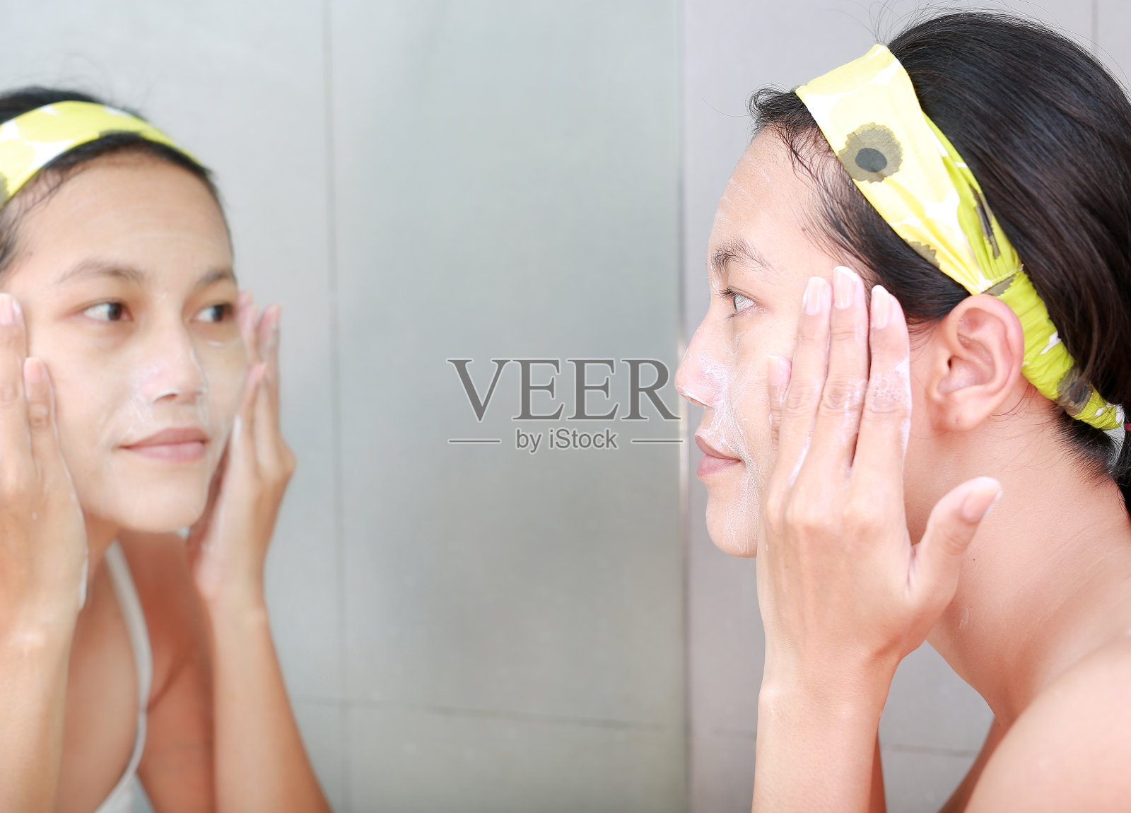 女人用浴室镜子在脸上涂抹泡沫霜。照片摄影图片