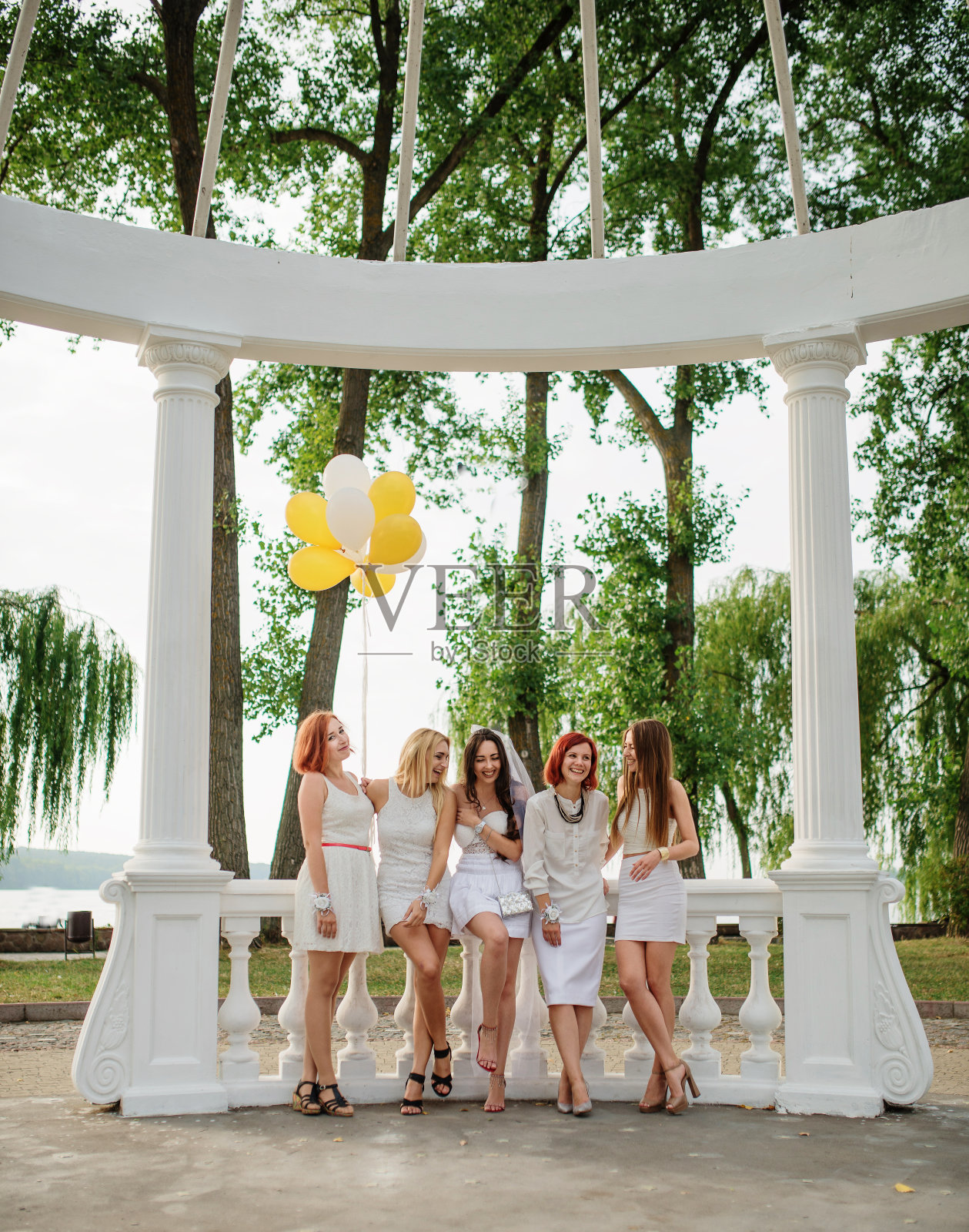 五个手里拿着气球的女孩穿着白色的衣服，在白色的拱门柱旁参加女性聚会。照片摄影图片