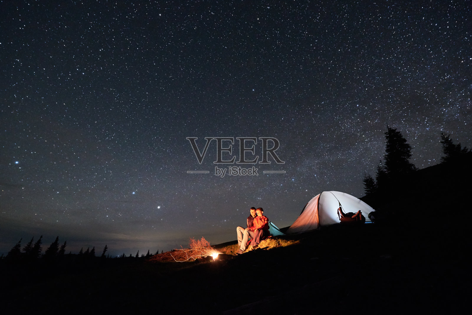 晚上露营。在美丽的夜空下，浪漫的游客夫妇在明亮的帐篷旁的篝火旁休息。天体摄影术插画图片素材