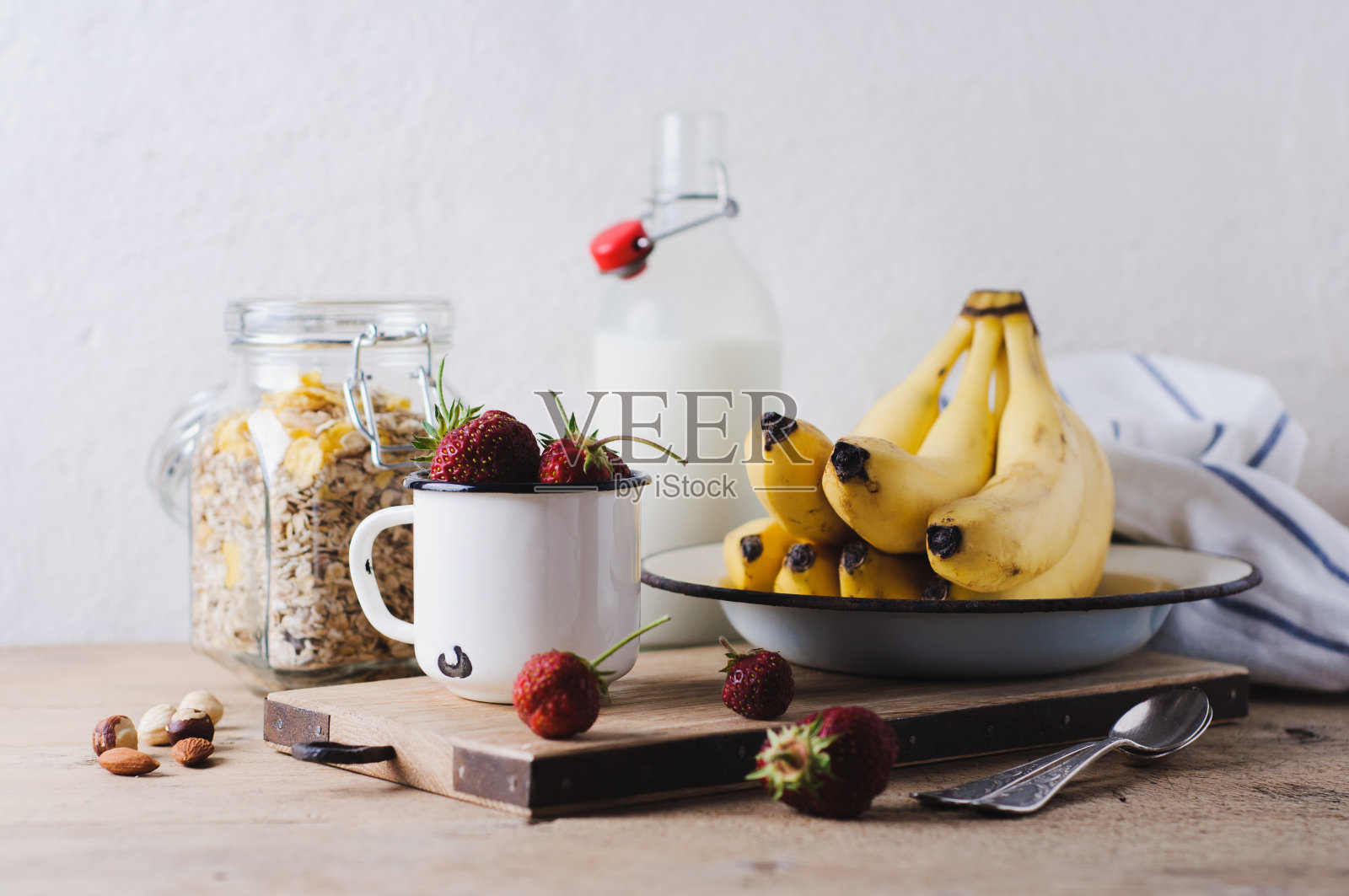 新鲜的草莓放在一个白色的金属杯里，香蕉放在砧板上，谷物和一瓶牛奶放在旧木桌上。早餐还是清洁饮食的概念照片摄影图片