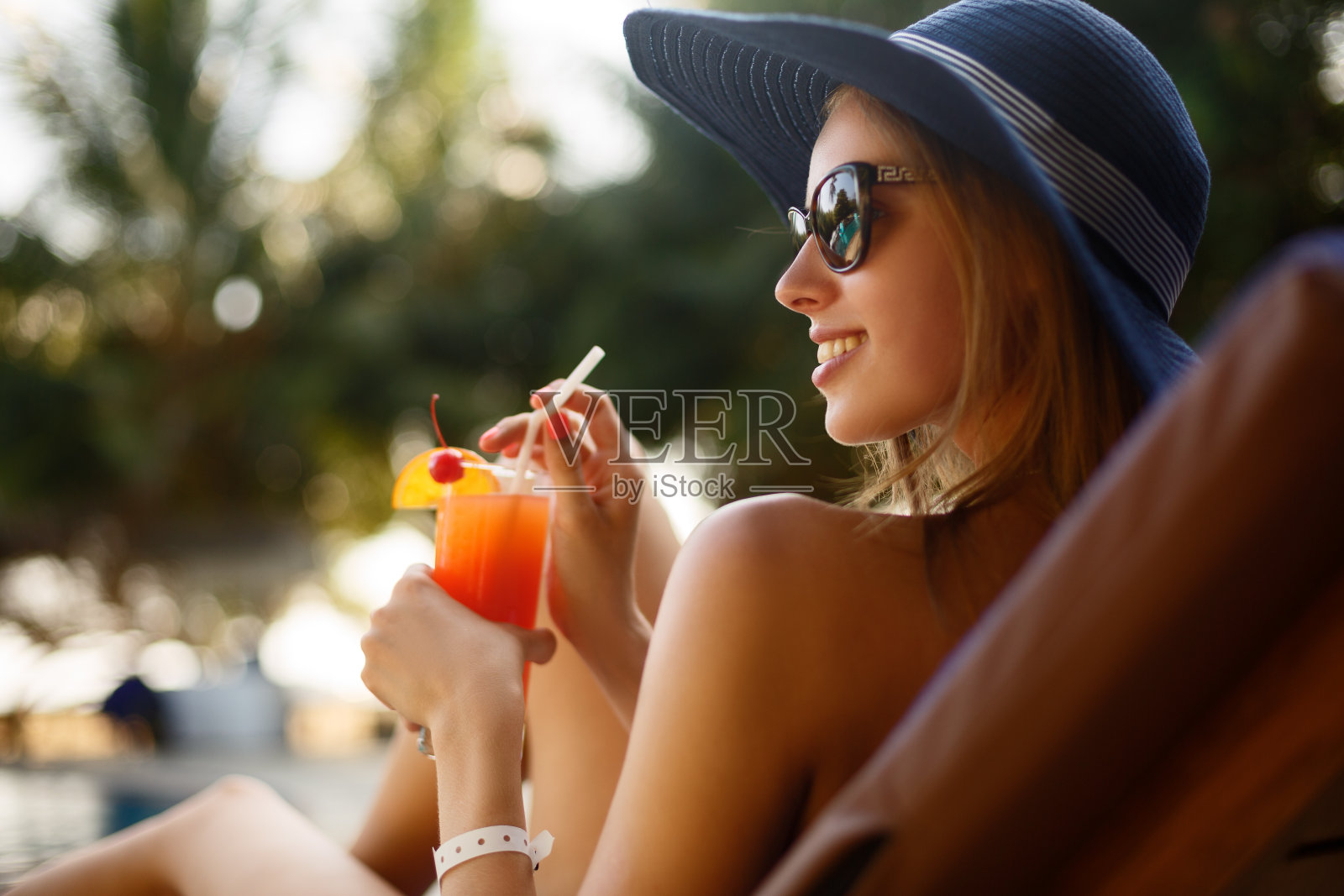 一个年轻女人的肖像与鸡尾酒杯在热带阳光下寒冷在游泳池附近的躺椅上，后面有棕榈树。假期的概念照片摄影图片