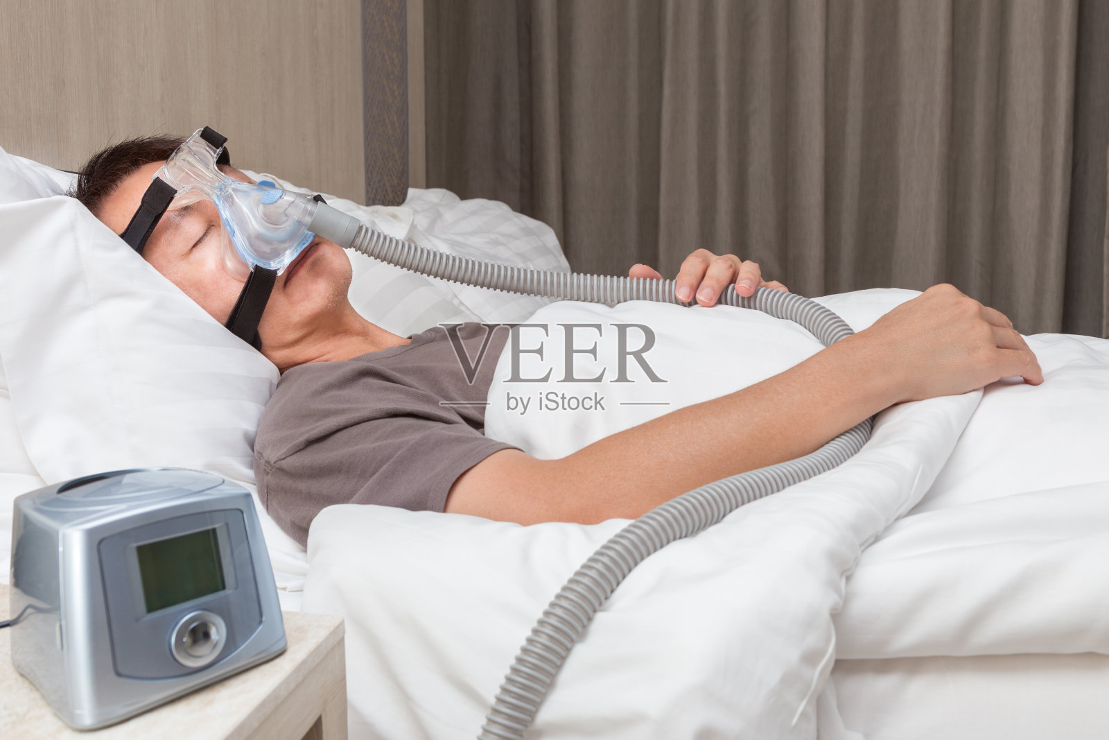 中年亚洲男子睡在床上，戴着连接呼吸软管的呼吸机面罩，床侧有呼吸机，用于治疗睡眠呼吸暂停症照片摄影图片