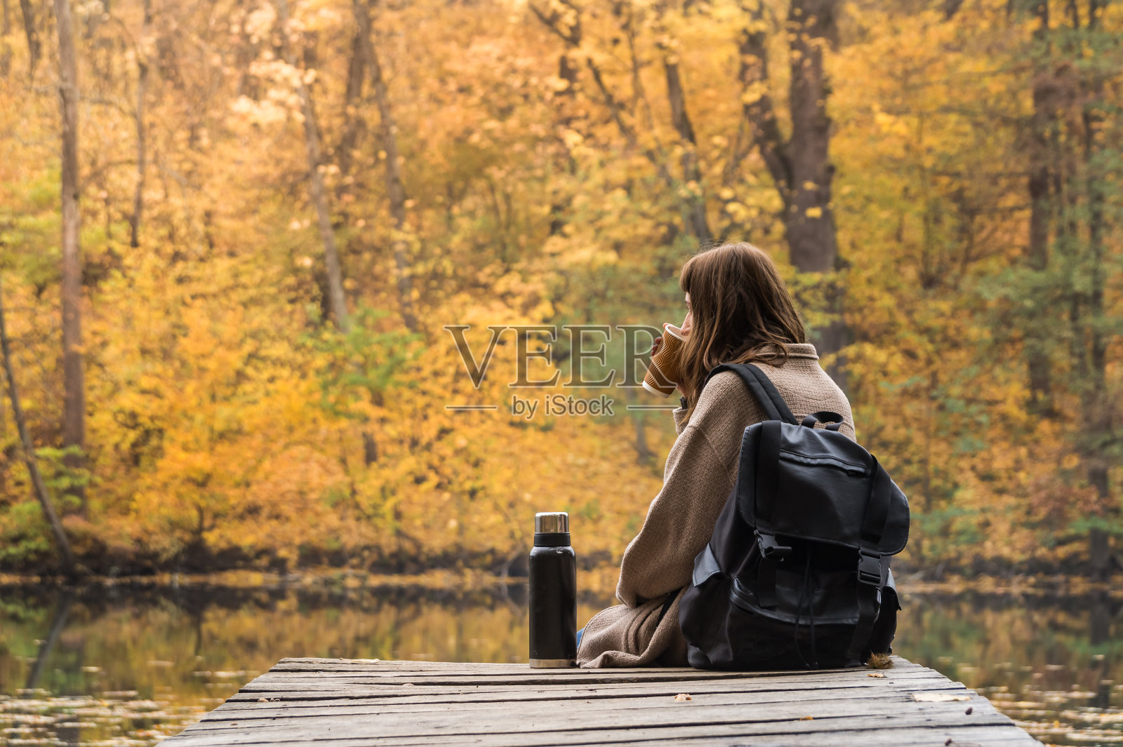 年轻的女徒步旅行者背包坐在河边，喝着咖啡，看着美丽的印度夏天的风景照片摄影图片