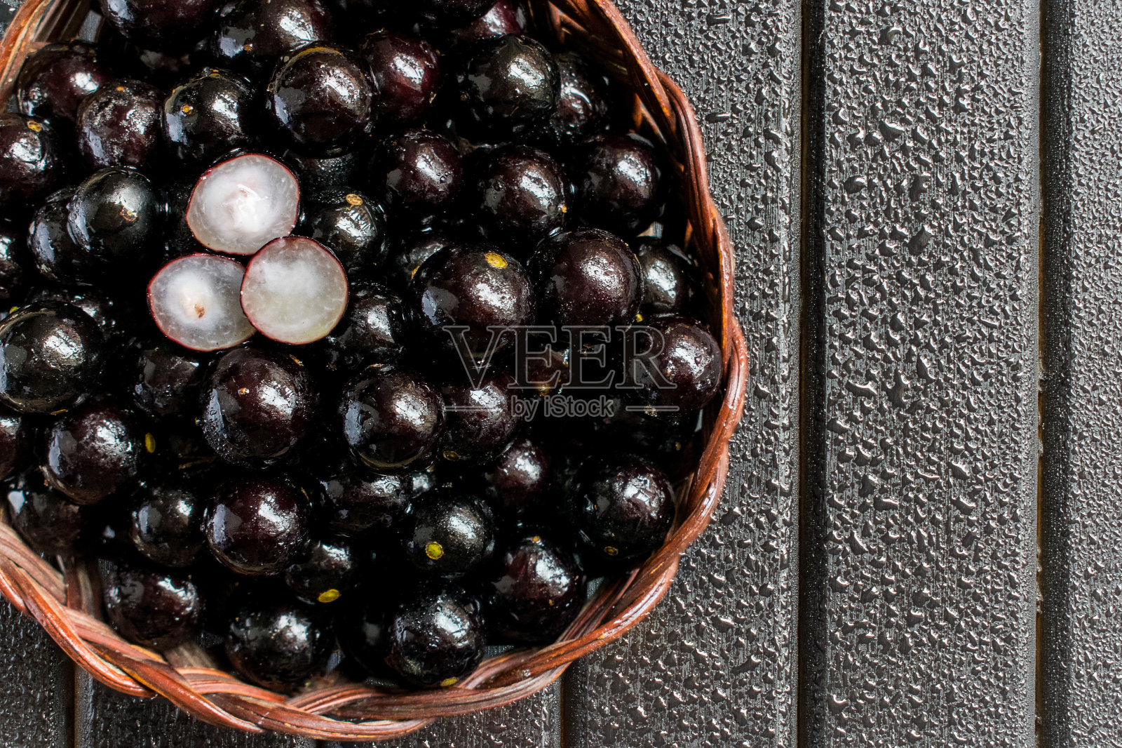Jabuticabas，也被称为巴西浆果或巴西葡萄藤(Plinia cauliflora)的果实放在一个木制的藤蔓篮子里，黑色的木质背景被水滴湿了照片摄影图片