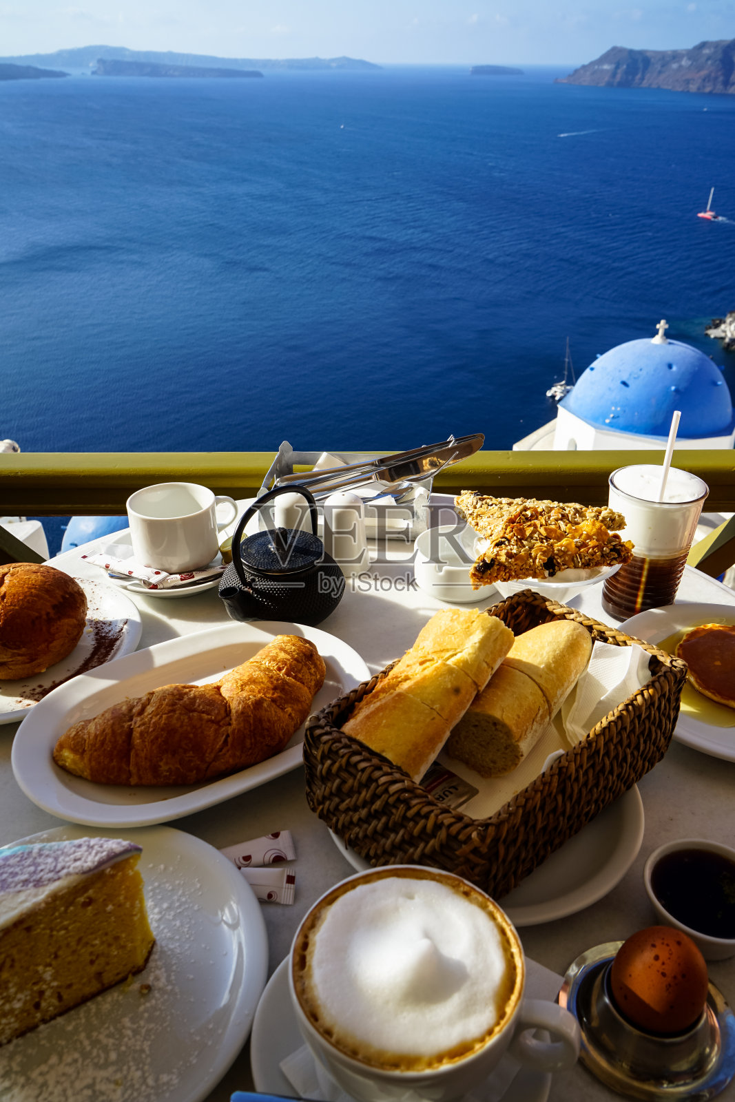 享受美丽的爱琴海海景早餐和伊亚教堂包括卡布奇诺，蛋糕，法式面包，羊角面包，煮鸡蛋，热茶，煎饼，谷物棒和面包在白色大理石桌上照片摄影图片