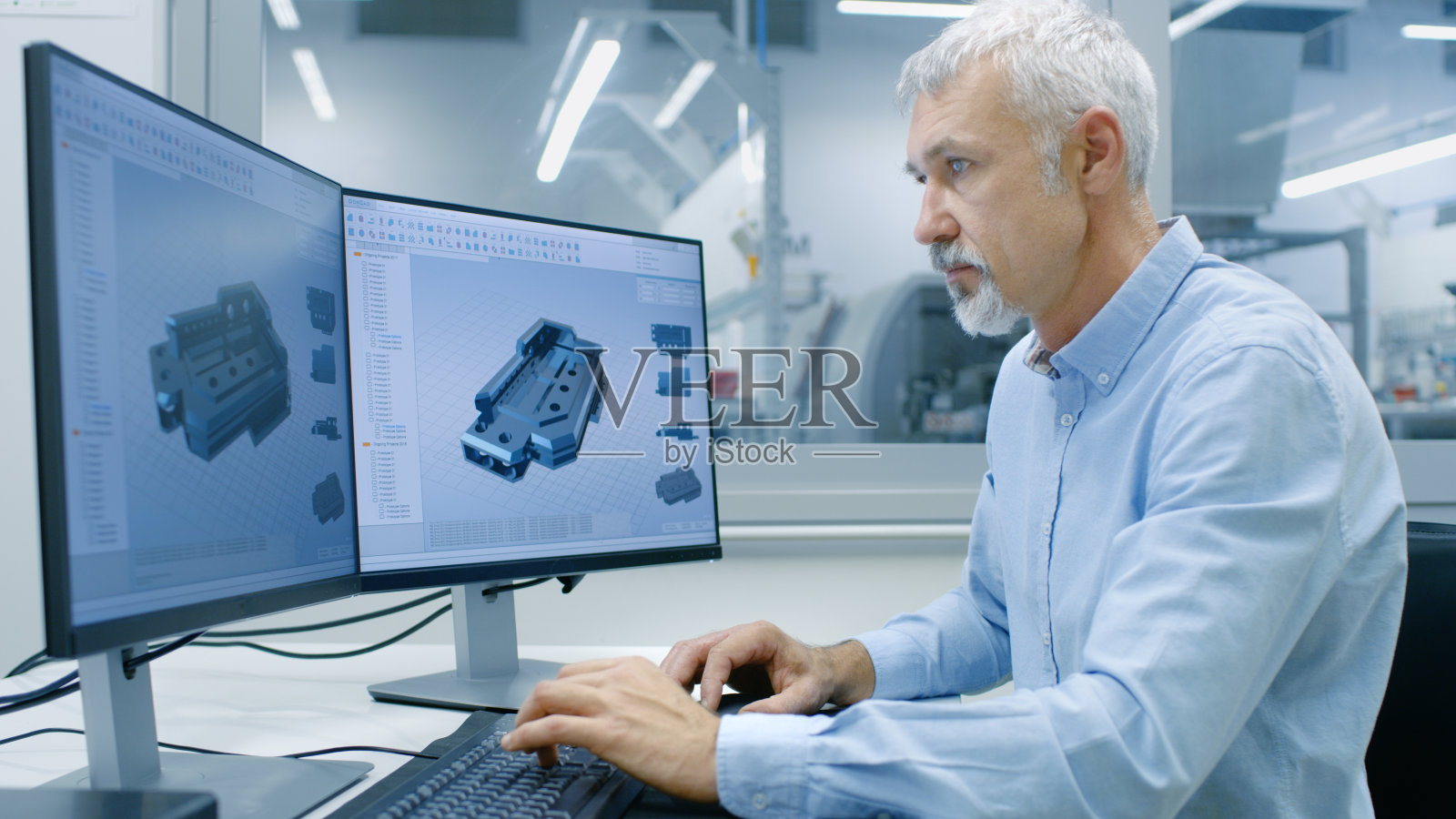 工程师工作在一个个人电脑与两个显示，他正在设计新的必要的CAD程序组件。可见办公室外窗口组件制造工厂。照片摄影图片