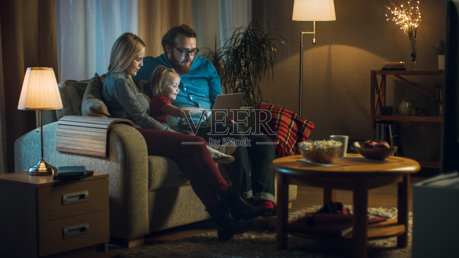 父亲，母亲和小女孩看电视的可能性不大。他们坐在舒适的客厅里的沙发上，父亲把笔记本电脑放在膝盖上。这是晚上。照片摄影图片