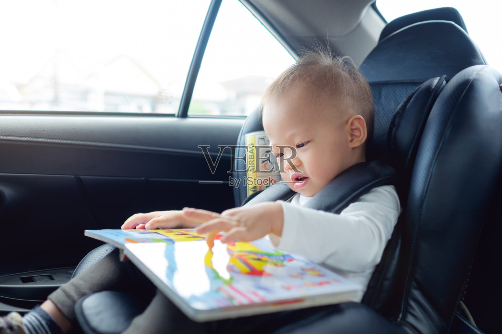 可爱的小亚洲18个月/ 1岁的蹒跚学步的小男孩坐在汽车座椅上，抱着，喜欢读书照片摄影图片