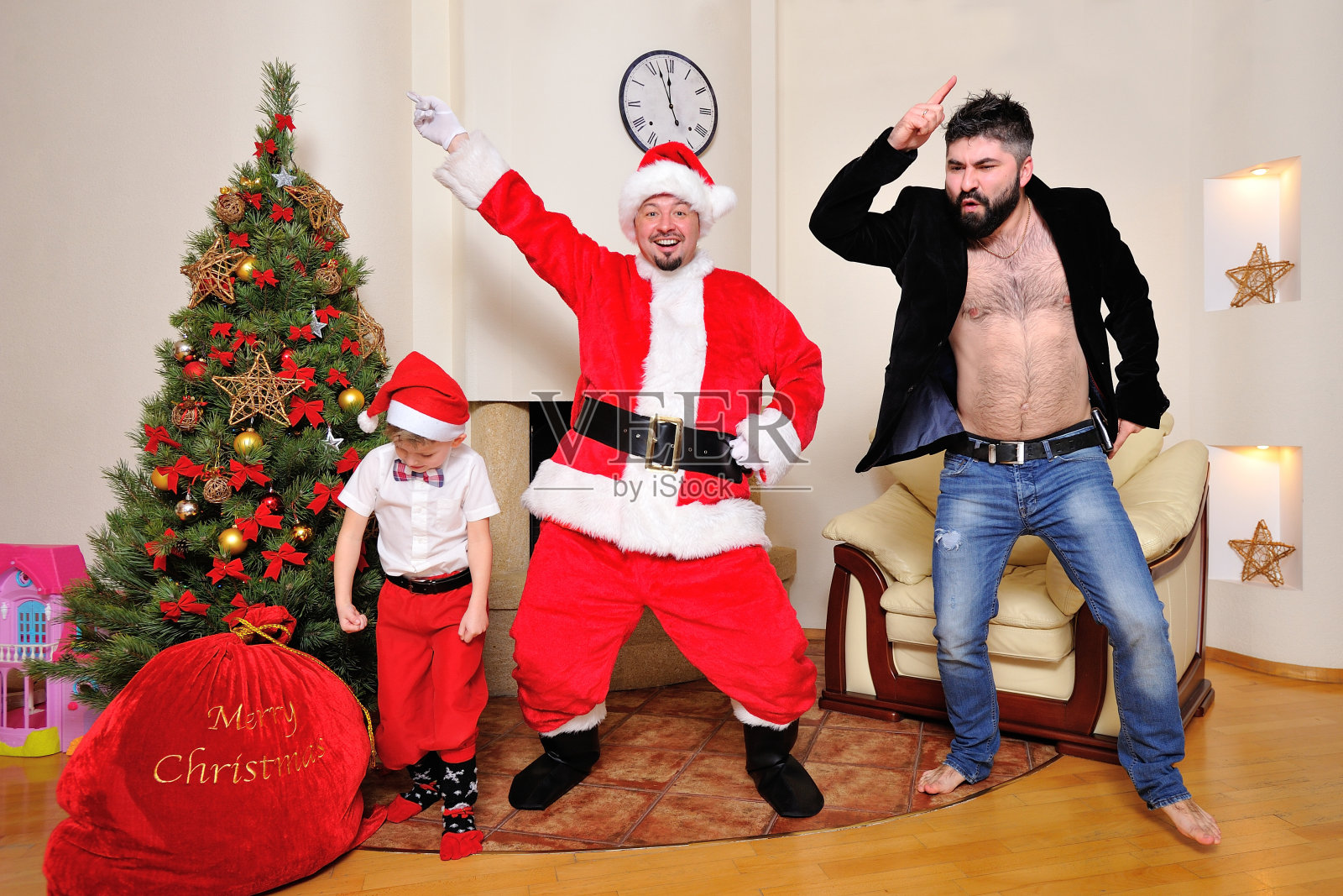 圣诞树，礼物袋，壁炉-男人打扮成圣诞老人，男孩在红色的裤子和高袜子和潮人在天鹅绒夹克裸露的上身跳舞，装傻。照片摄影图片