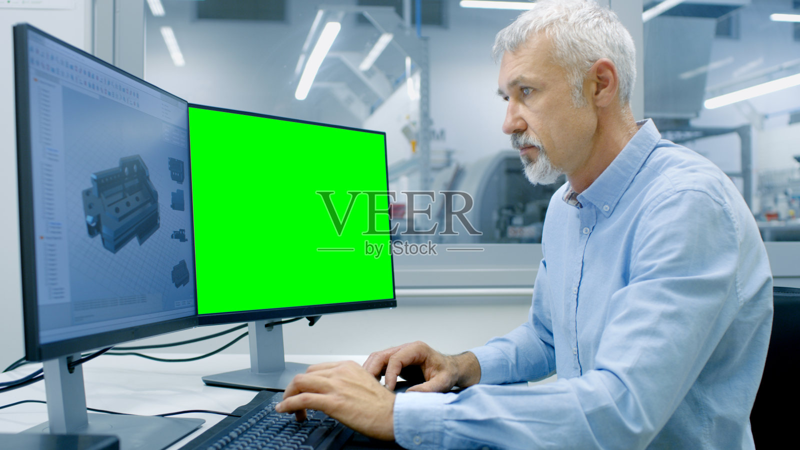 工程师工作在一个有两个显示器的计算机上，一个有色度键伟大的模型模板和CAD程序中的其他组件。可见办公室外窗口组件制造工厂。照片摄影图片