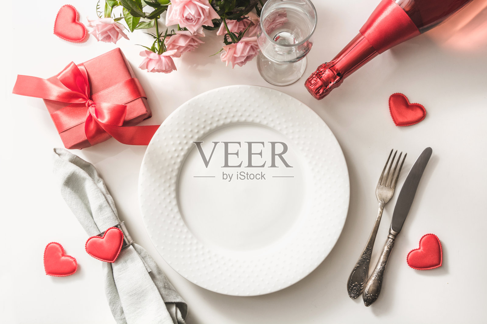 情人节晚餐与桌子的位置设置红色礼物，玻璃为香槟，一瓶香槟，粉红玫瑰，心形饰品与银器在白色的背景。前视图。情人节卡片。照片摄影图片