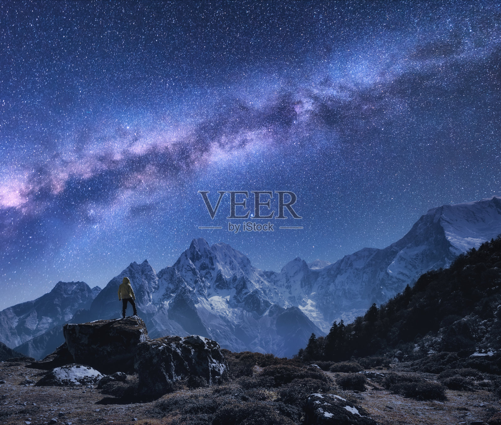 有银河和山脉的空间。在尼泊尔的夜晚，站在石头上、山上、星空上的人。白雪皑皑的山峰映衬着满天繁星。徒步旅行。夜景与明亮的银河照片摄影图片