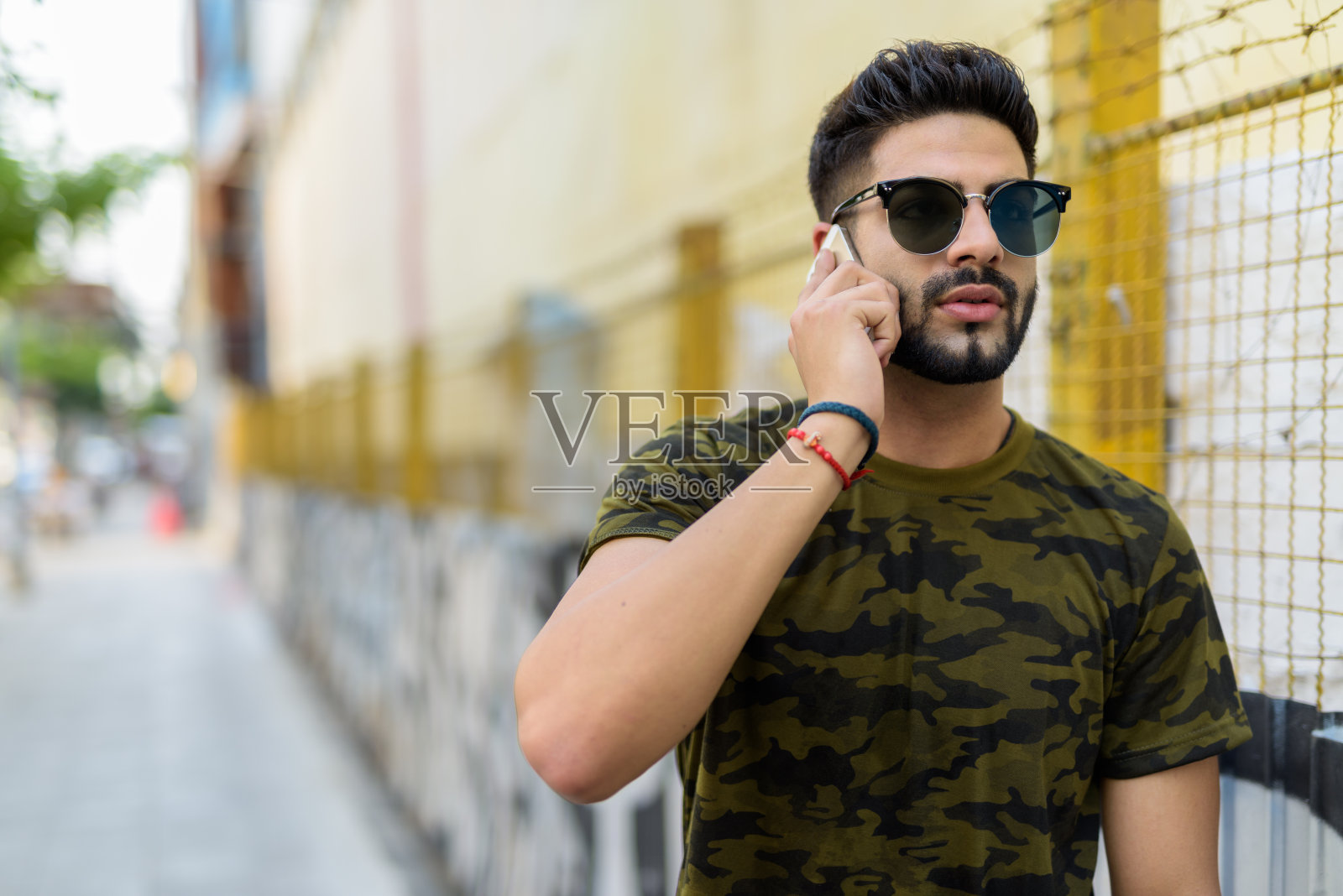 年轻英俊的印度男子戴着太阳镜，一边思考一边用手机说话，对着户外公共街道上涂鸦的肮脏的城市墙壁照片摄影图片