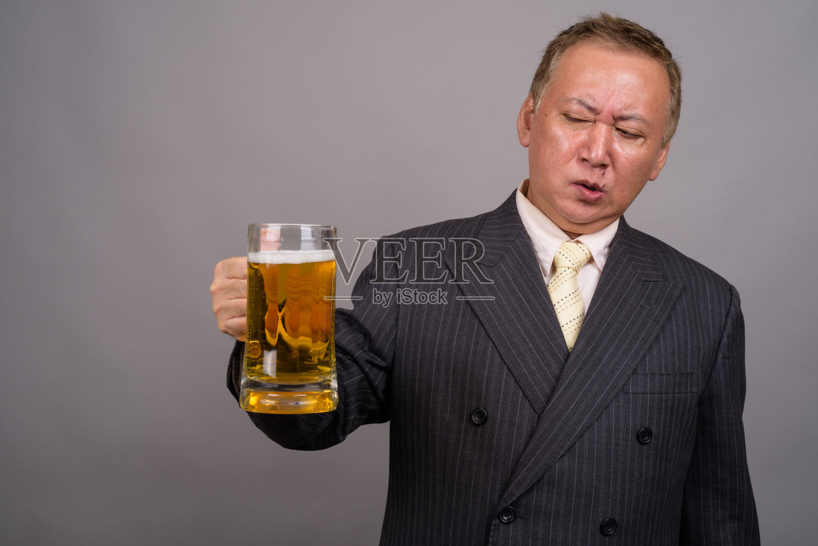 在灰色的背景下，一个成熟的亚洲商人拿着一杯啤酒照片摄影图片