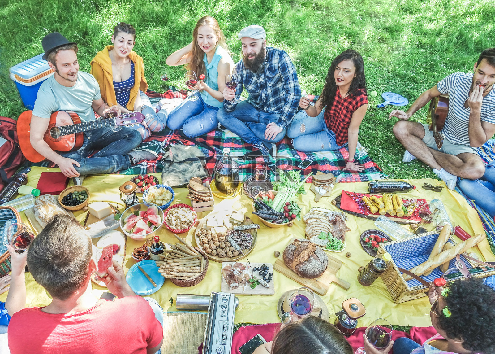 半平的观点，朋友在自然公园野餐坐在草地上-年轻人玩音乐，吃和喝烧烤派对-主要集中在中心顶部的家伙-对比过滤器照片摄影图片