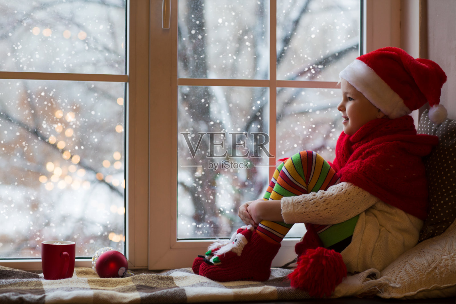 可爱的小女孩坐在窗边看雪。房间在圣诞节装饰。照片摄影图片