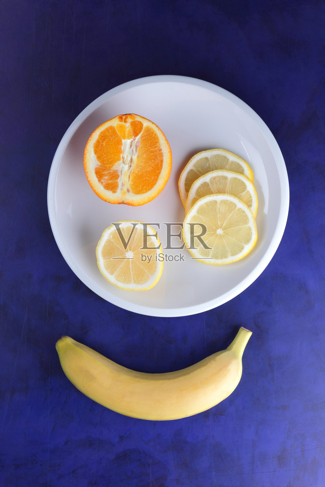 极简主义风格的紫外光背景柑橘，白色盘子上的柠檬片和半片橙子，深蓝色背景上的黄色香蕉，健康食品，素食，流行艺术照片摄影图片
