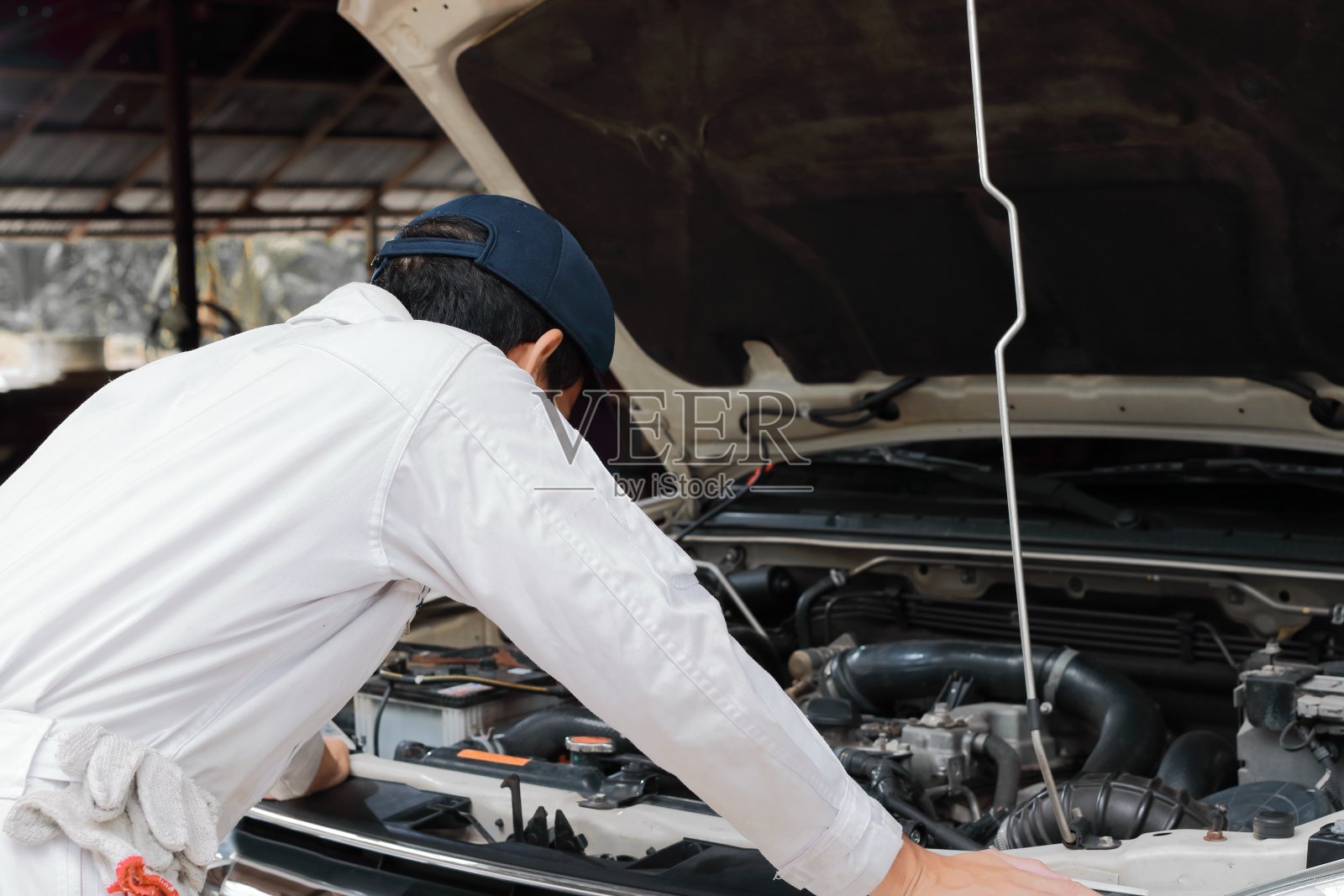 沮丧压力年轻的机修工在白色制服感到失望或疲惫与汽车在开放的引擎盖在修理厂。照片摄影图片