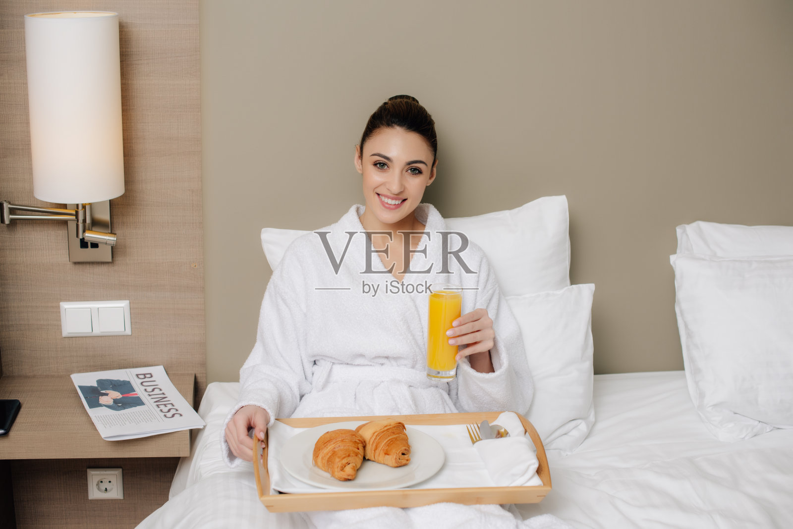 微笑的女人在浴衣放松在酒店房间的床上早餐和看相机照片摄影图片