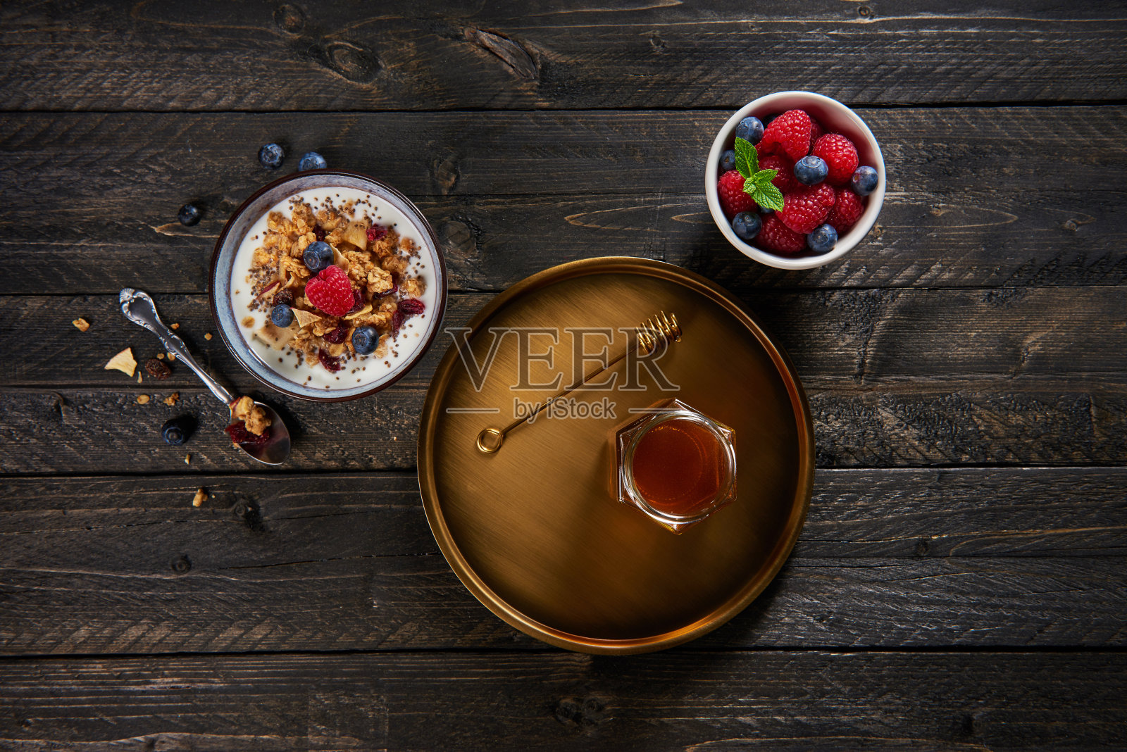 俯视图的健康早餐。牛奶什锦早餐，酸奶，蓝莓和覆盆子在碗和枫糖浆在金盘。深色木制背景。照片摄影图片