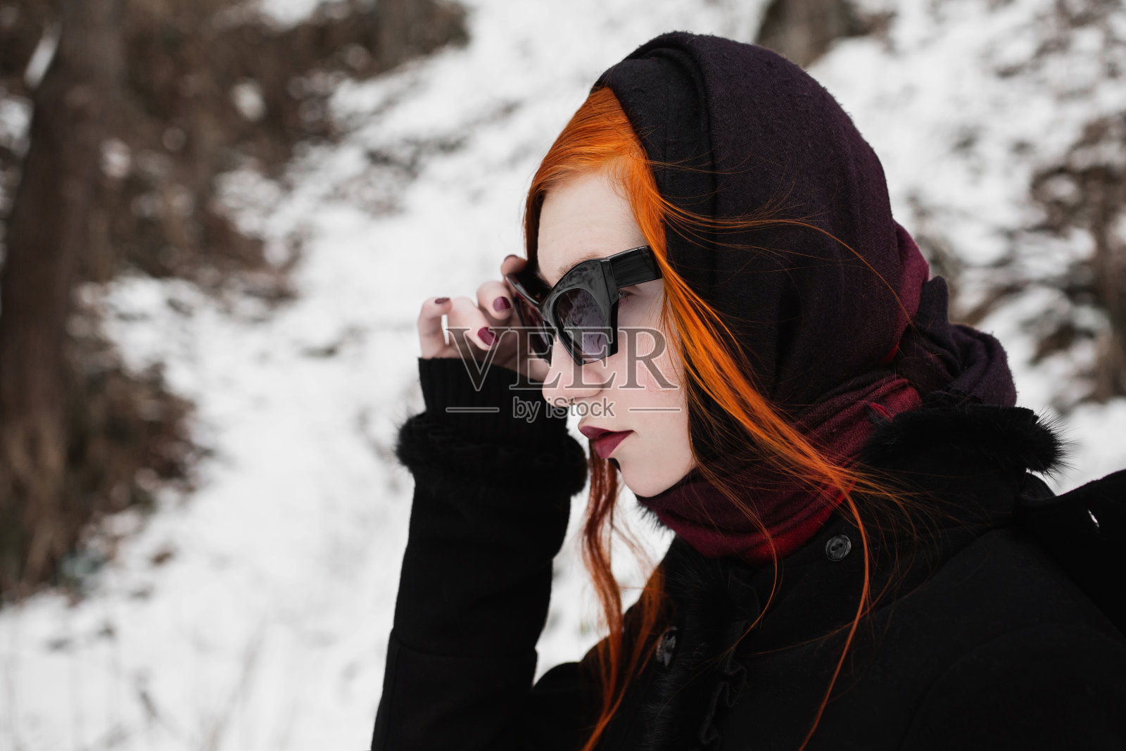 红发时髦的女孩，苍白的皮肤，蓝色的眼睛，酒红色的嘴唇，她的头披着一条围巾，穿着一件黑色的外套，在一个冬天的森林背景，女人与黑色太阳镜肖像的侧面照片摄影图片