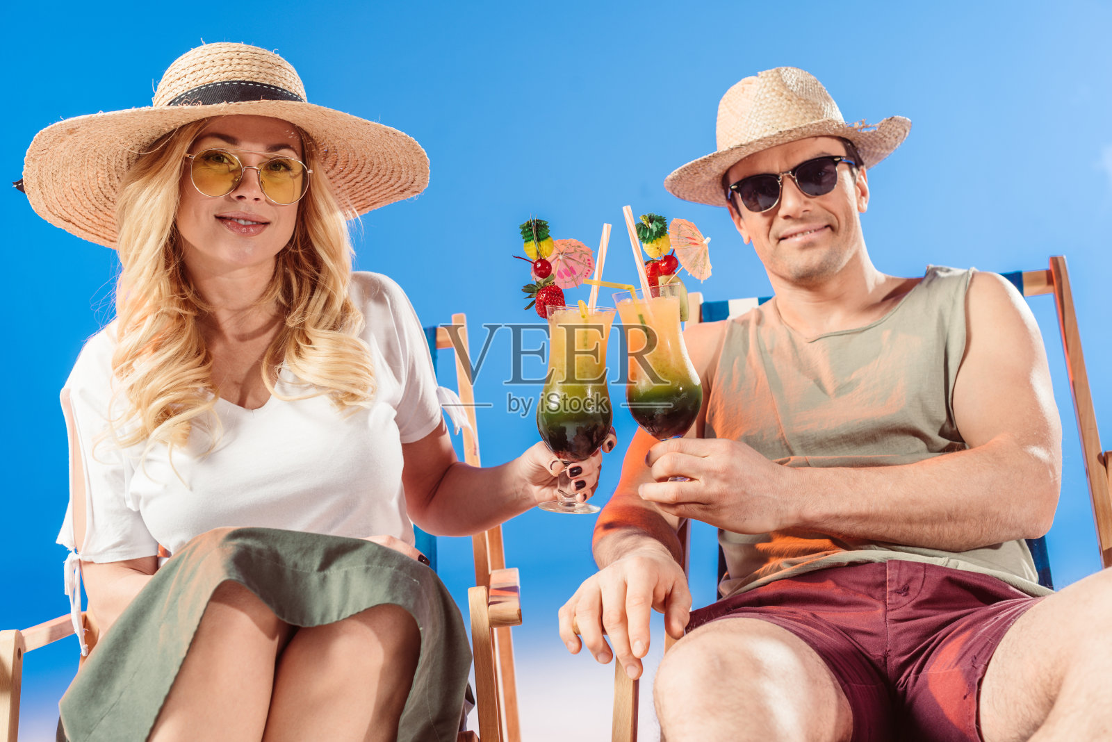一对年轻迷人的夫妇坐在蓝色背景的躺椅上喝着鸡尾酒敬酒照片摄影图片