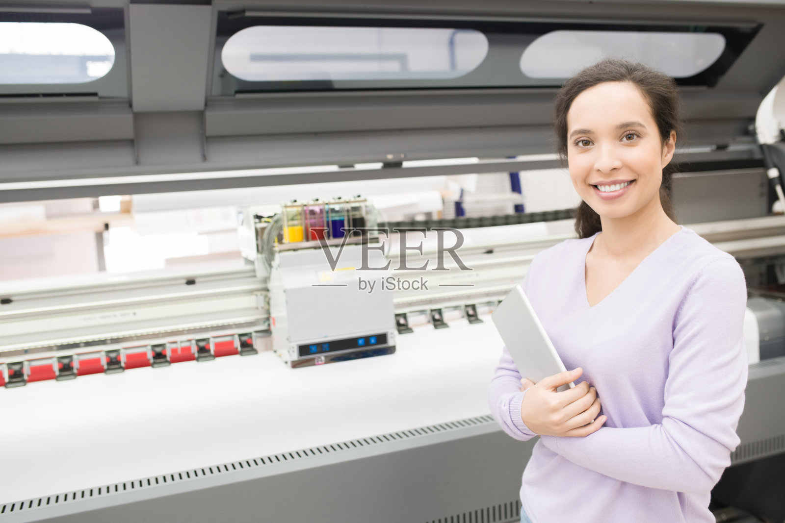 快乐自信的女性打印操作员拿着平板电脑，看着相机愉快地站在大版面打印机照片摄影图片