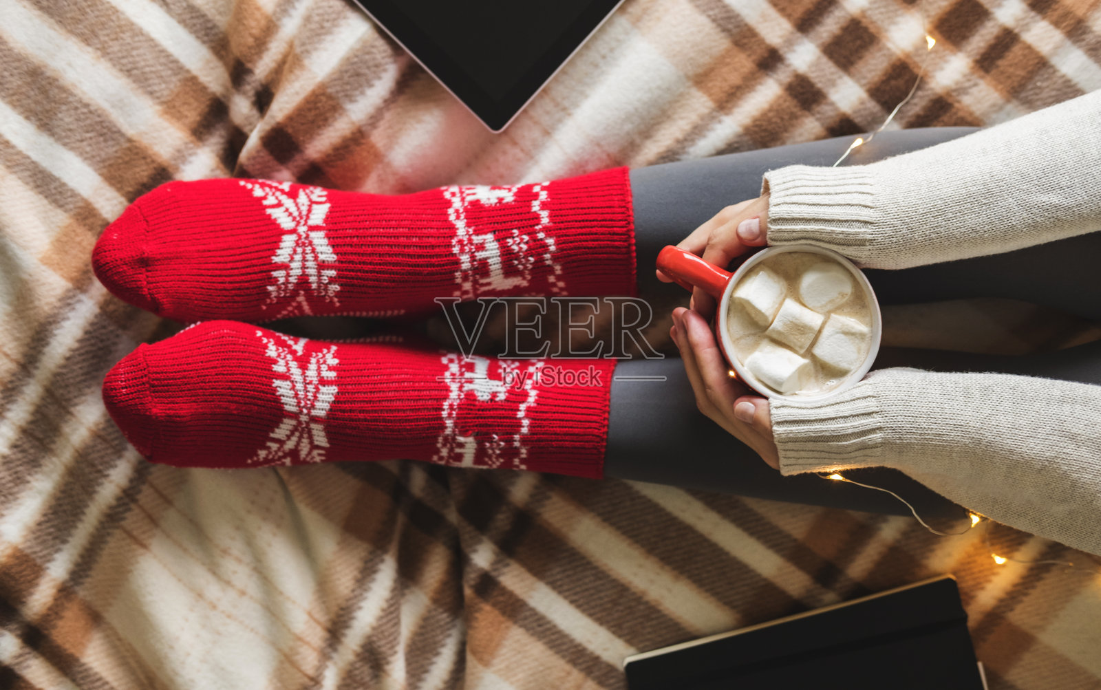 女人的手和脚穿着毛衣和舒适的红色羊毛袜子拿着一杯热咖啡和棉花糖，坐在格子花环，平板电脑和笔记本。概念冬季舒适，清晨畅饮。照片摄影图片