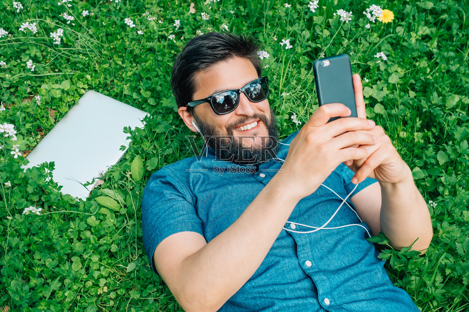 一个留着胡子的年轻人的肖像，他正放松地躺在草地上，用着智能手机照片摄影图片