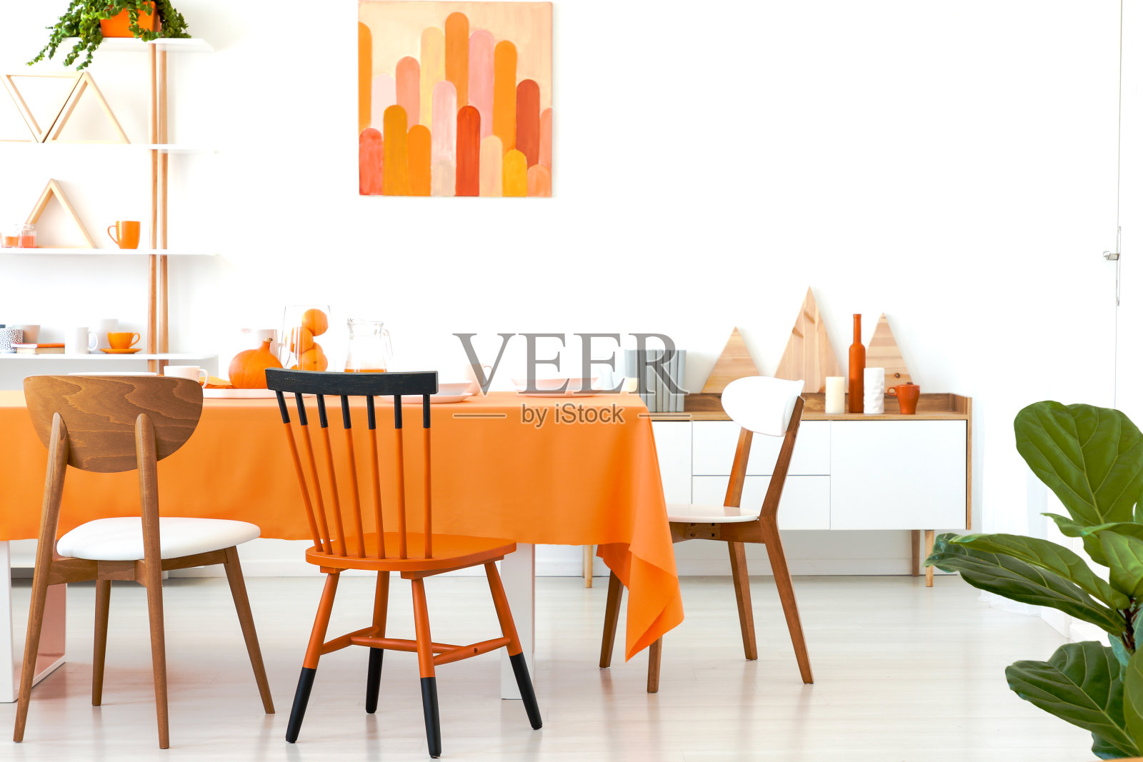 三个不同的椅子放置在长餐桌与橙色桌布和餐具在真实的照片白色的房间内部与装饰架，简单的海报和橱柜与书照片摄影图片