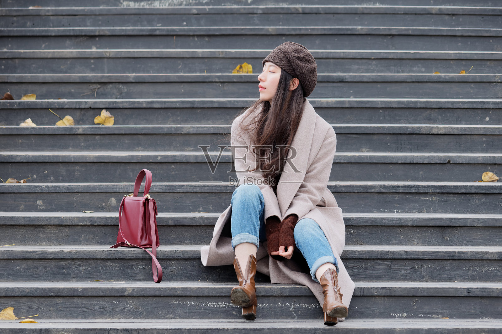 肖像美丽的年轻女子看远思考坐在一个公园的木制台阶在户外的背景深秋，与红色的袋子在她旁边。照片摄影图片