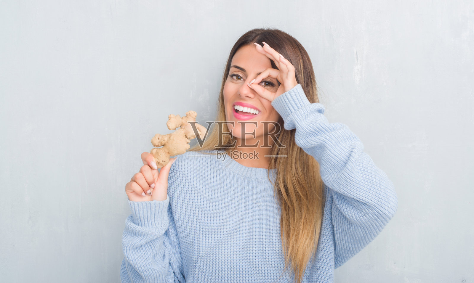 年轻的成年妇女在灰色的垃圾墙抱着新鲜的生姜蔬菜与快乐的脸微笑做ok手势与手在眼睛透过手指照片摄影图片