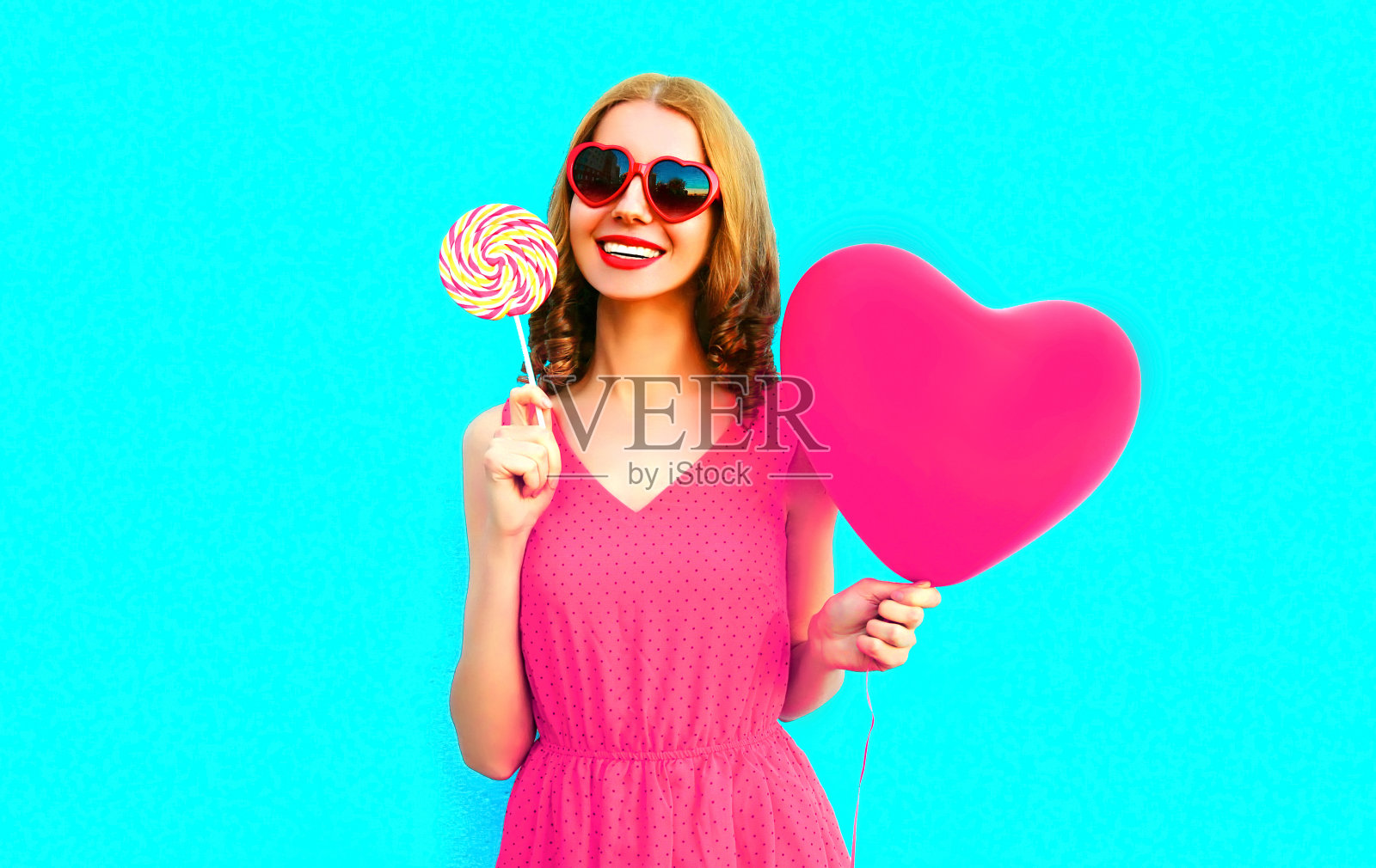 肖像快乐微笑的年轻女子与棒棒糖，粉红色的心形气球在彩色的蓝色背景照片摄影图片