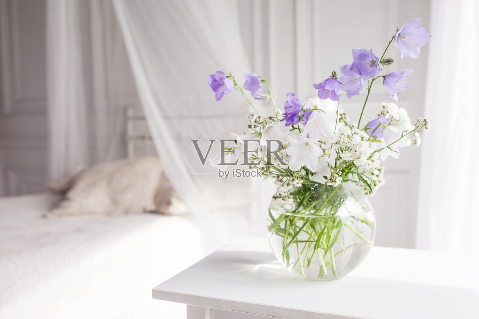 淡紫色和白色花朵的玻璃花瓶在明亮舒适的卧室内部。白色的墙壁，白色的床单，浅色的毯子或格子和枕头。照片摄影图片
