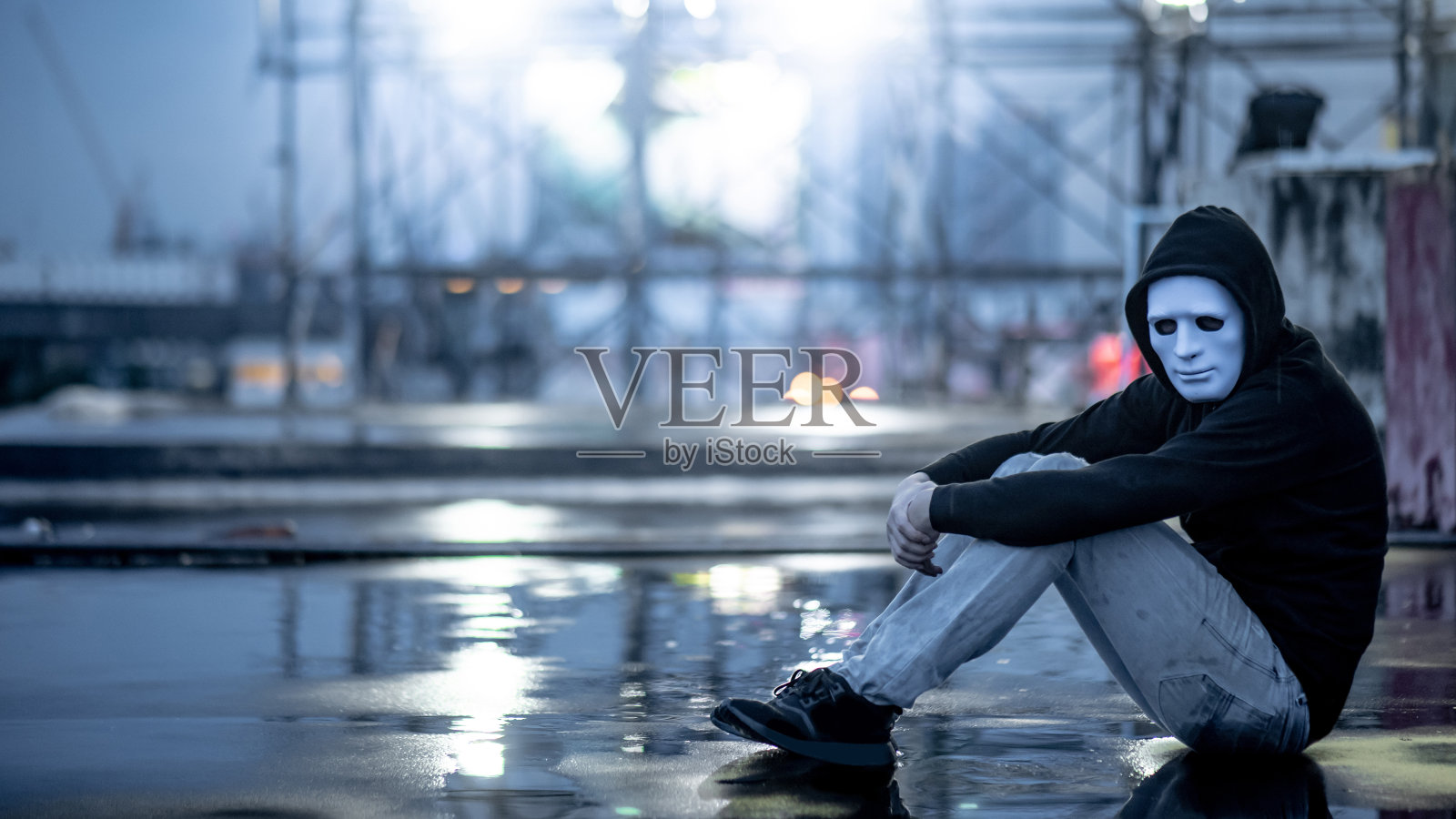 这是一个穿着白色面罩的神秘帽衫男子抱着膝盖坐在废弃建筑的屋顶上淋雨的画面。双相情感障碍或重度抑郁症。抑郁症的概念照片摄影图片