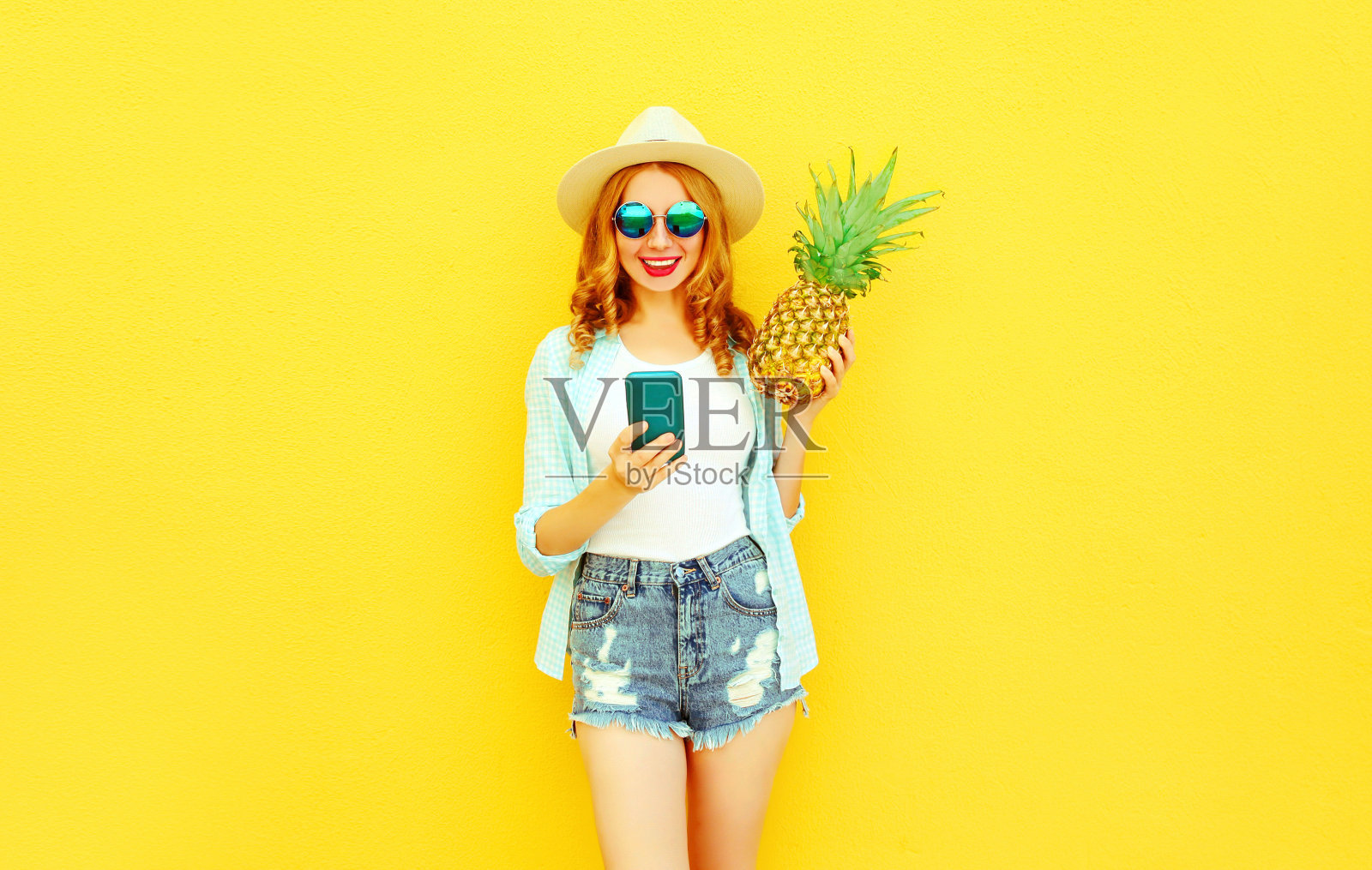 快乐微笑的女人拿着智能手机，菠萝在夏天草帽，短裤上彩色的黄色墙壁背景照片摄影图片