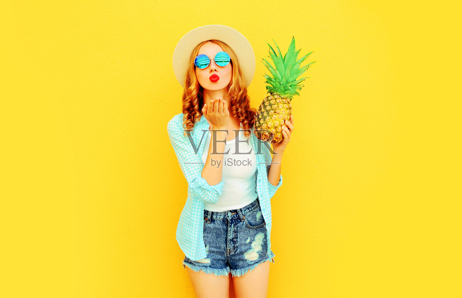 肖像女子与菠萝送甜蜜的飞吻在夏季草帽，太阳镜，短裤在彩色的黄色背景照片摄影图片