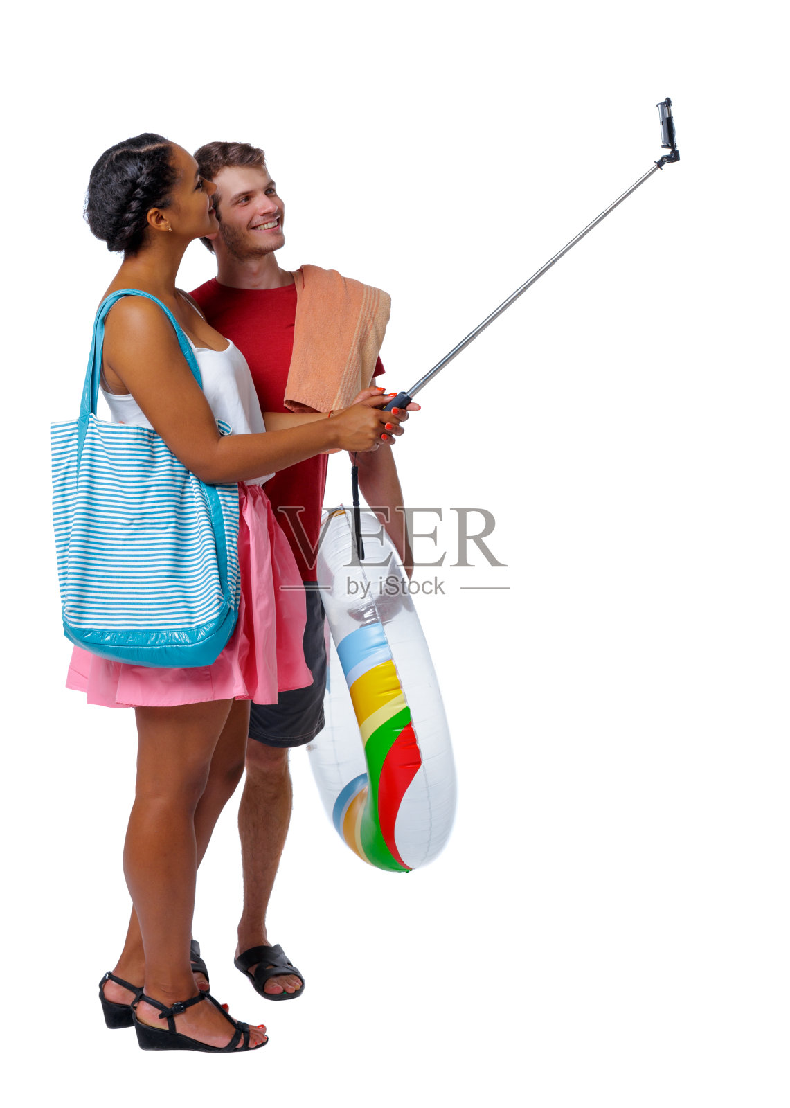 一对国际夫妇的侧视图，在自拍杆上用充气圈和沙滩配件自拍。照片摄影图片