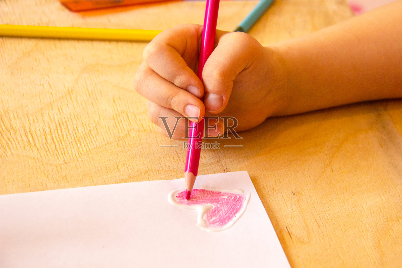 孩子用铅笔在白纸上画，背景是木制的纹理。艺术品工作场所与创意配件。特写的孩子的手画一个粉红色的心照片摄影图片