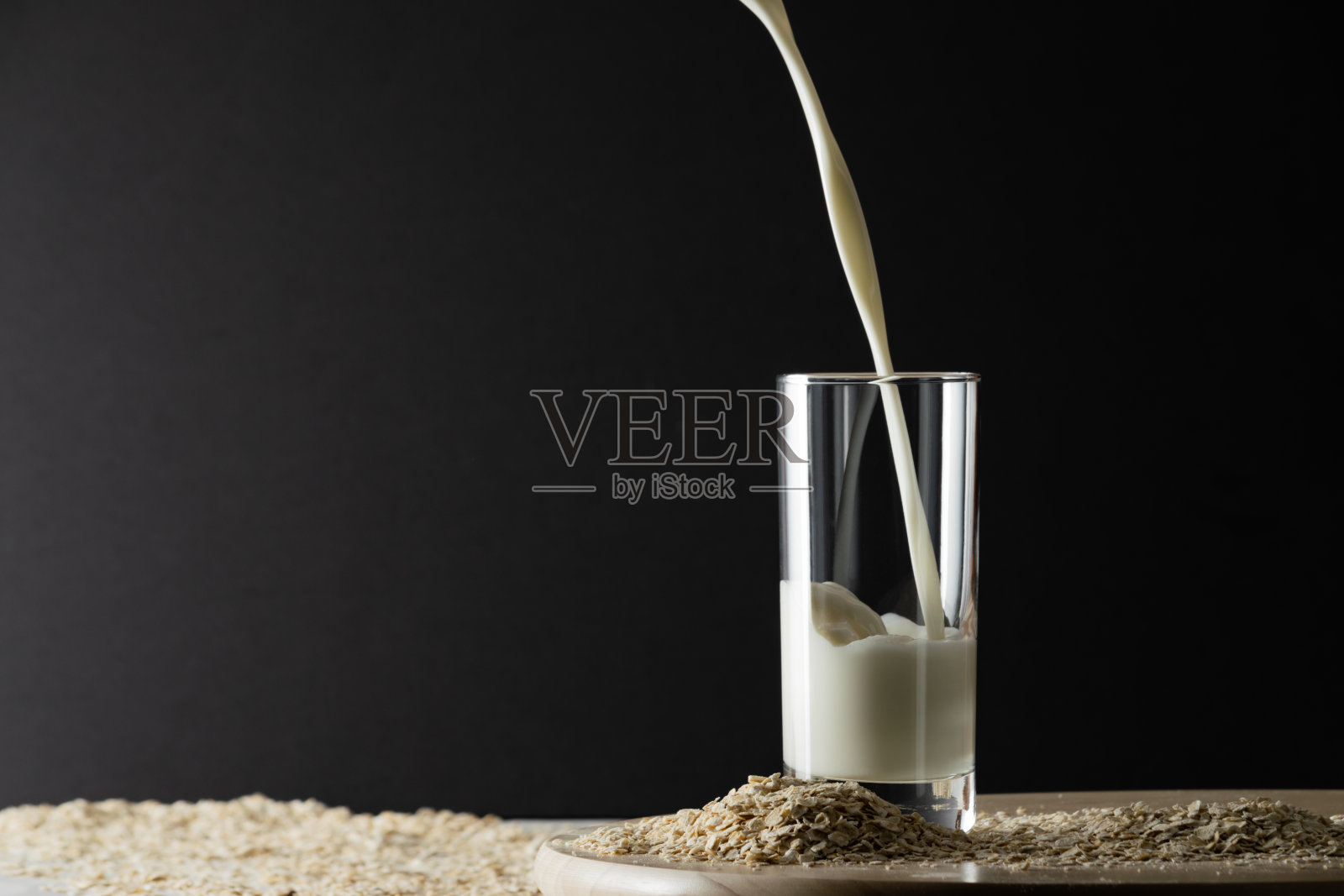 将燕麦牛奶倒入黑色背景的玻璃杯中，将燕麦片放在桌上。无乳糖的燕麦奶替代品概念。复制空间，柔和的阴影。照片摄影图片