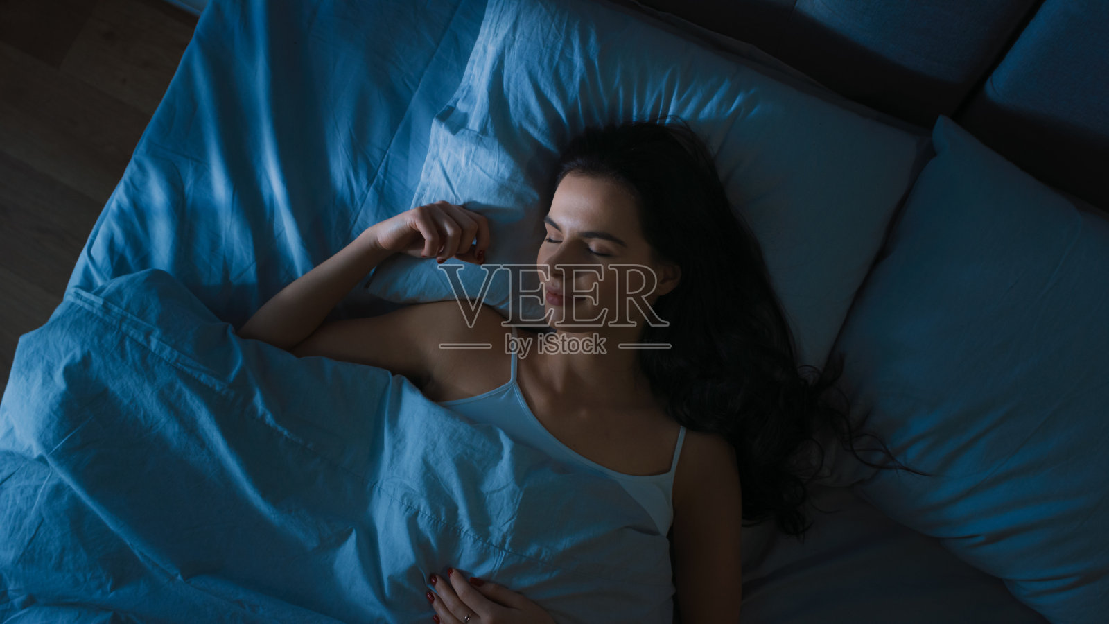 俯视图美丽的年轻女子舒适地睡在床上在他的卧室晚上。蓝色的夜间颜色和寒冷微弱的灯柱灯光透过窗户。照片摄影图片