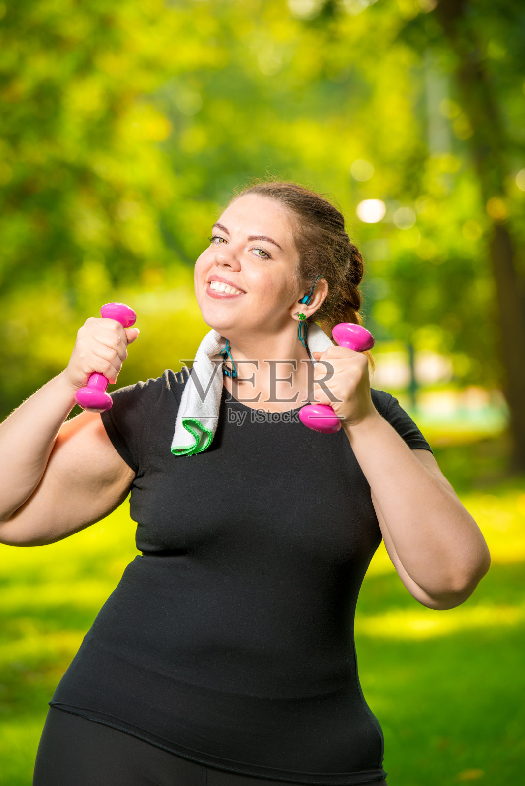 垂直肖像特大号妇女戴着耳机，手里拿着哑铃正在公园里进行运动照片摄影图片