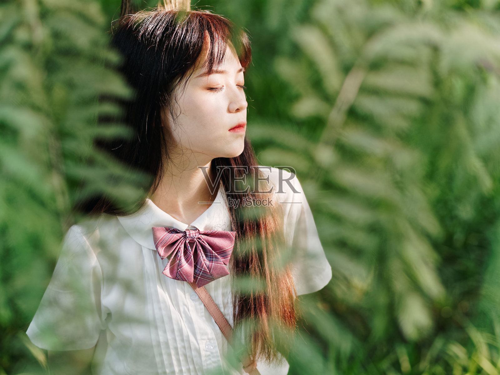 在阳光明媚的夏日森林里，美丽的中国女孩穿着白衬衫，靠在一棵树上，闭着眼睛呼吸着新鲜的空气。美丽，生活方式和夏季概念。照片摄影图片
