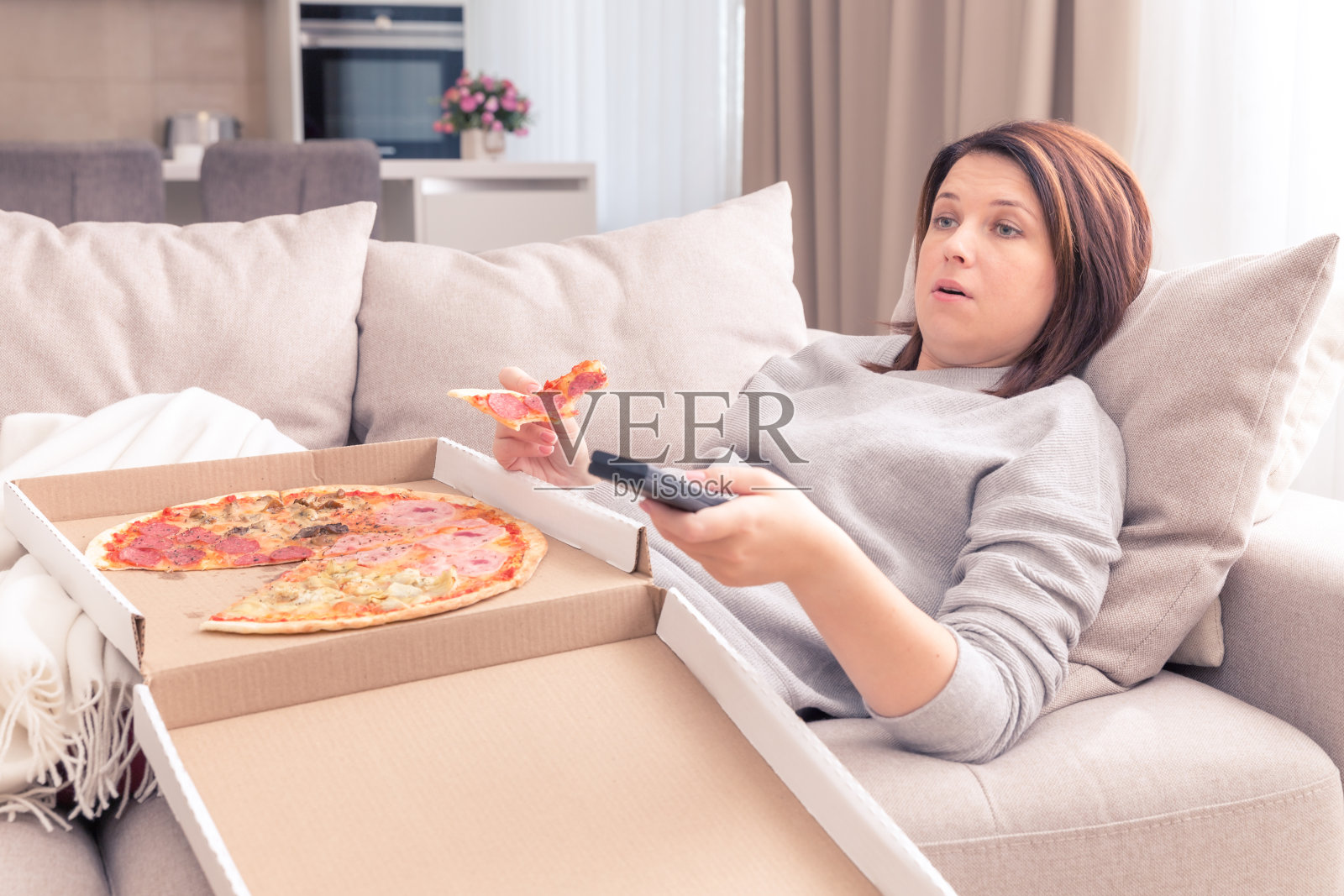 惊讶的女人在家里一边吃披萨一边用遥控器看电视，暖色调照片摄影图片