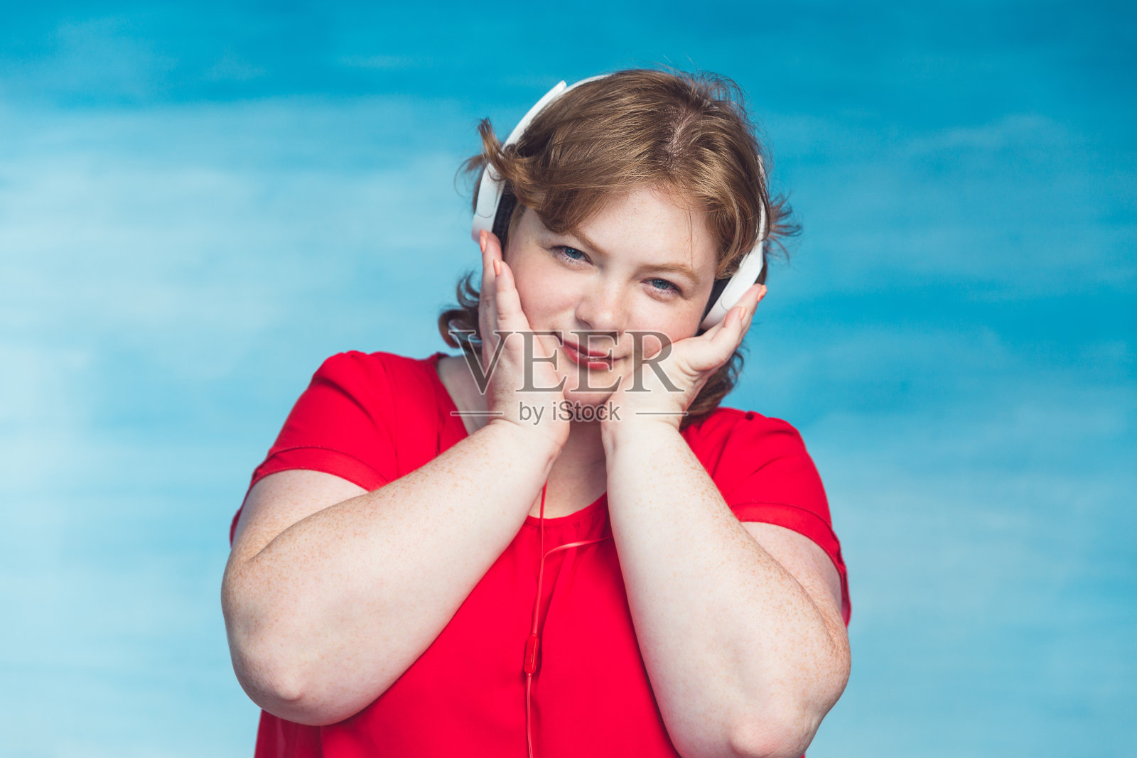 年轻迷人的红发胖女人戴着赏心悦目的耳机在蓝色背景下听音乐。照片摄影图片
