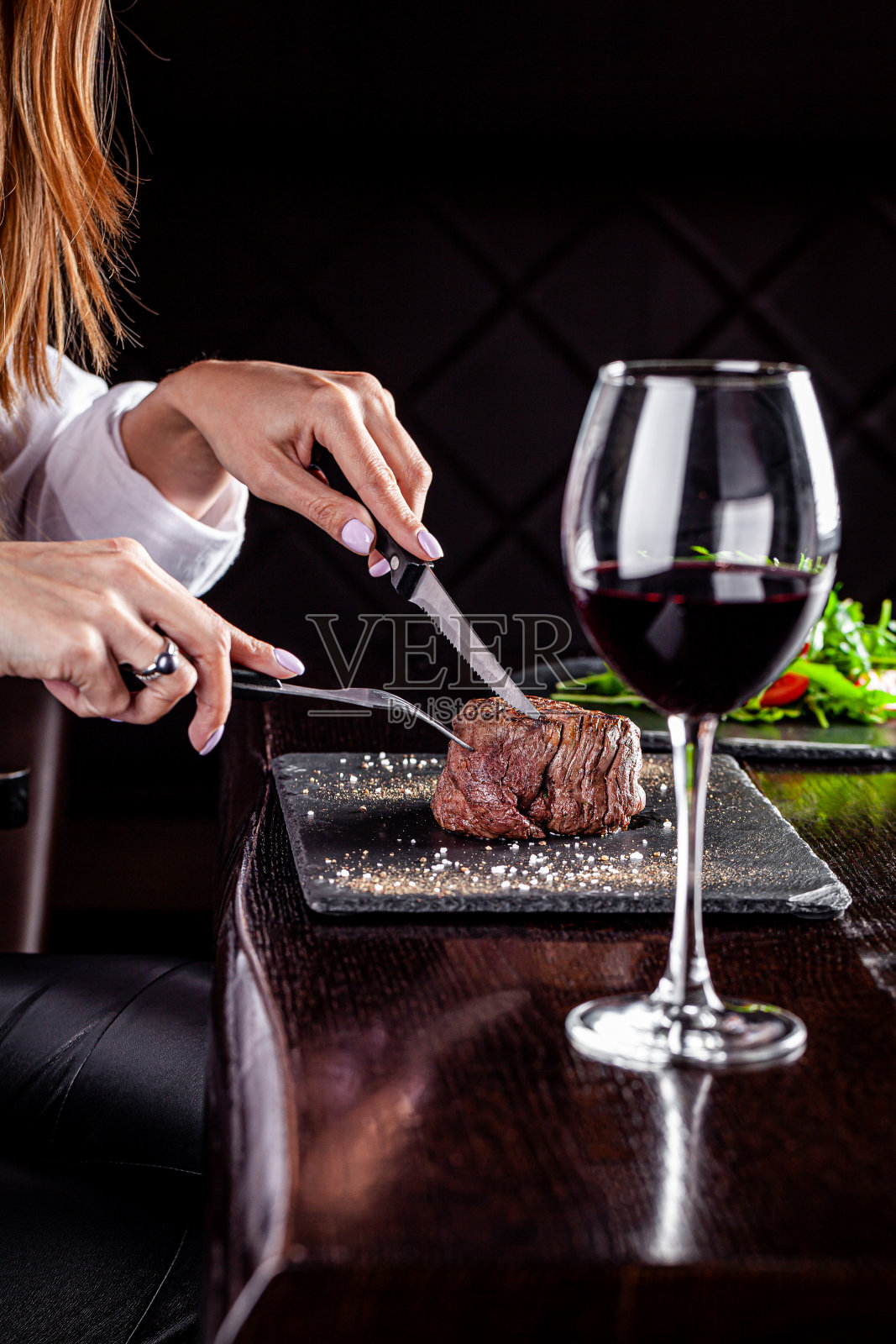 美国菜。餐馆里的那个女孩有一块多汁的牛排，手里拿着刀叉。桌子上有一杯红酒。背景图像，拷贝空间照片摄影图片