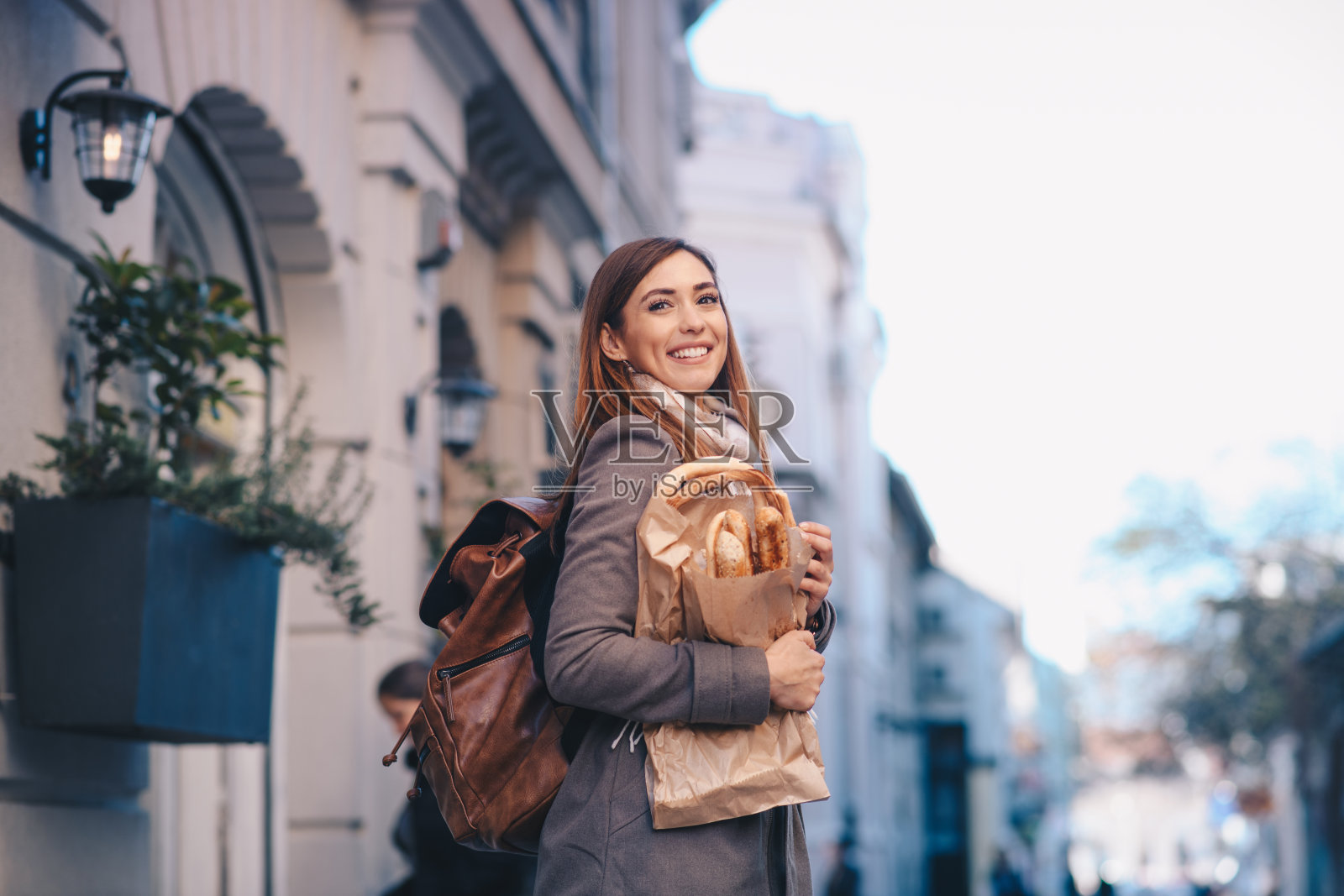 一个女人拿着一袋面包制品在街上。她肩上戴着另一个包。照片摄影图片