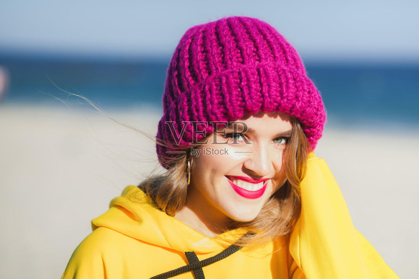 年轻漂亮的女人在黄色的运动衫和明亮的紫色针织帽在海滩上照片摄影图片