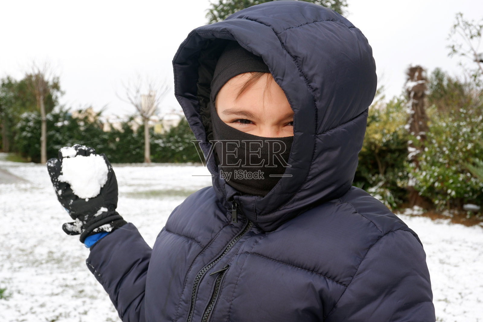 快乐的小男孩戴着雪面罩，右手拿着雪球。照片摄影图片