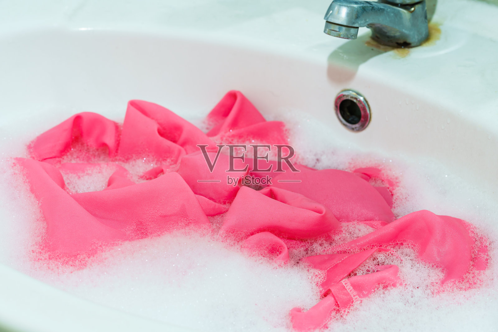 粉红色的织物被浸泡在洗涤槽的洗涤剂中。照片摄影图片